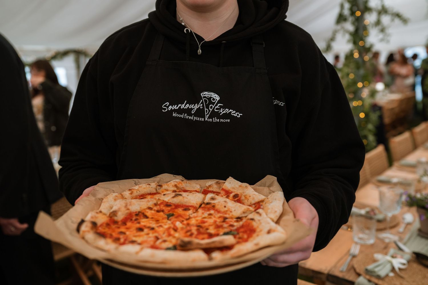 Someone in a black hoodie holds a freshly baked pizza on brown parchment paper at an event setting.
