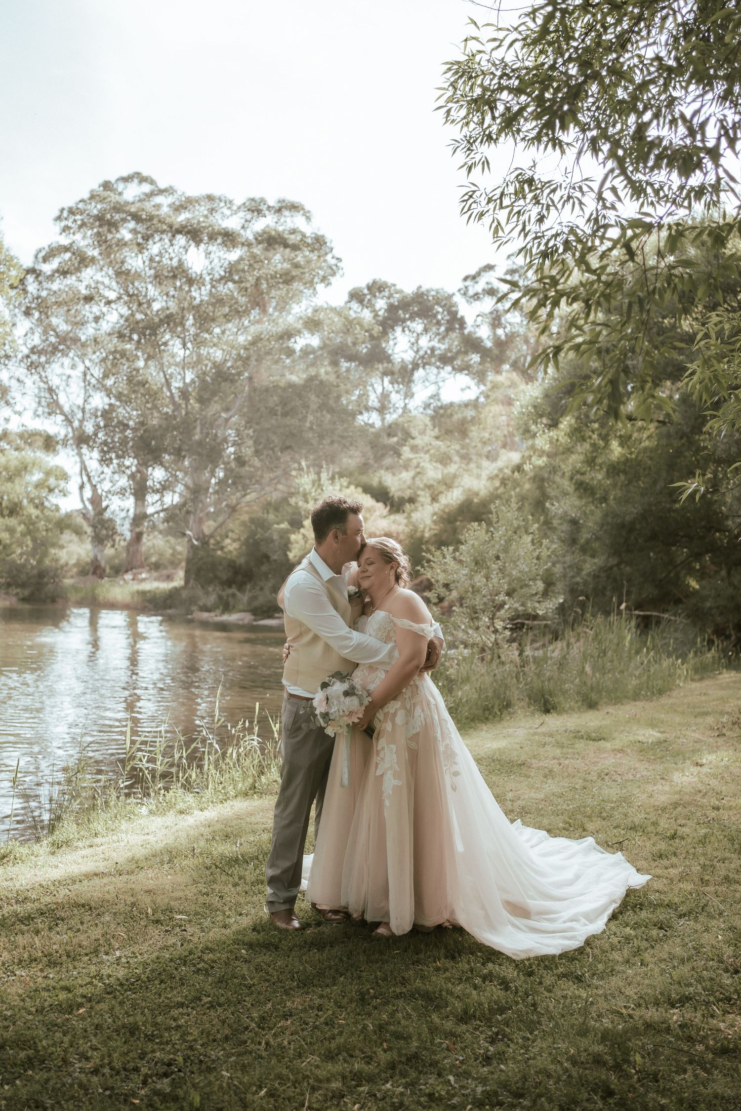 A couple embraces on their wedding day in an outdoor setting near a tranquil lake with eucalyptus trees in the background along Howqua River