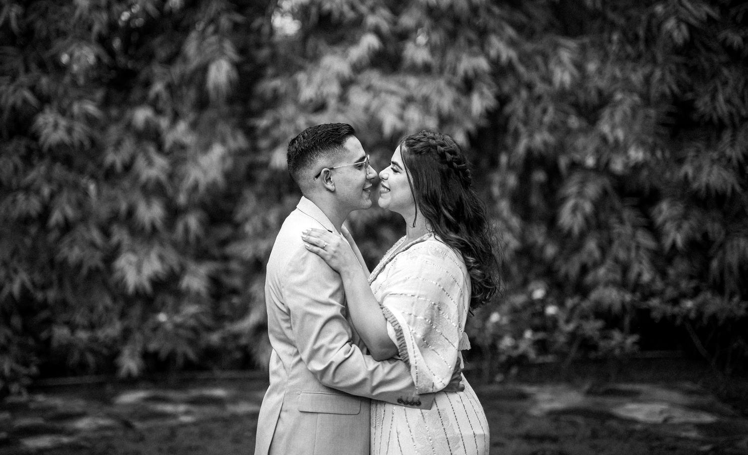 Black and white romantic photograph of a couple in a tender embrace surrounded by lush greenery in a garden setting.