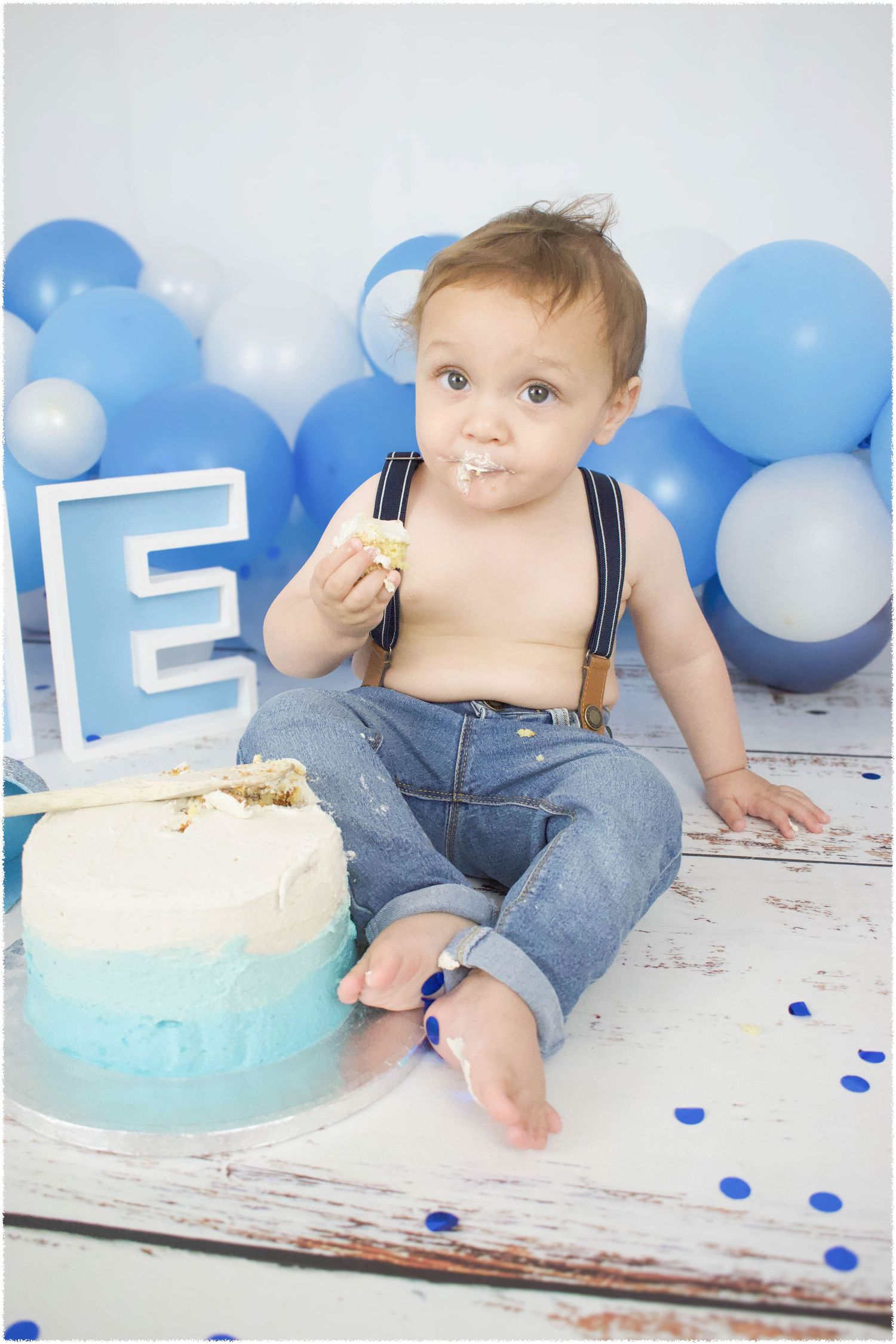Baby in denim overalls enjoys cake smash photo session with blue balloons.