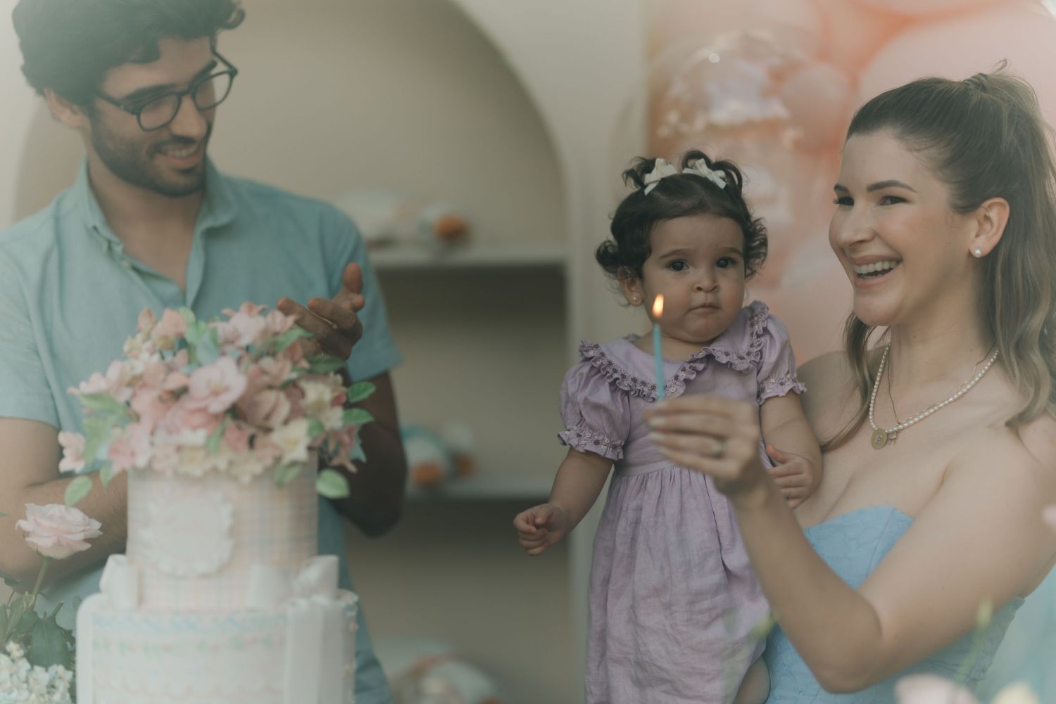 Group celebrates child's birthday around a pink floral cake at home with family and friends gathered together.