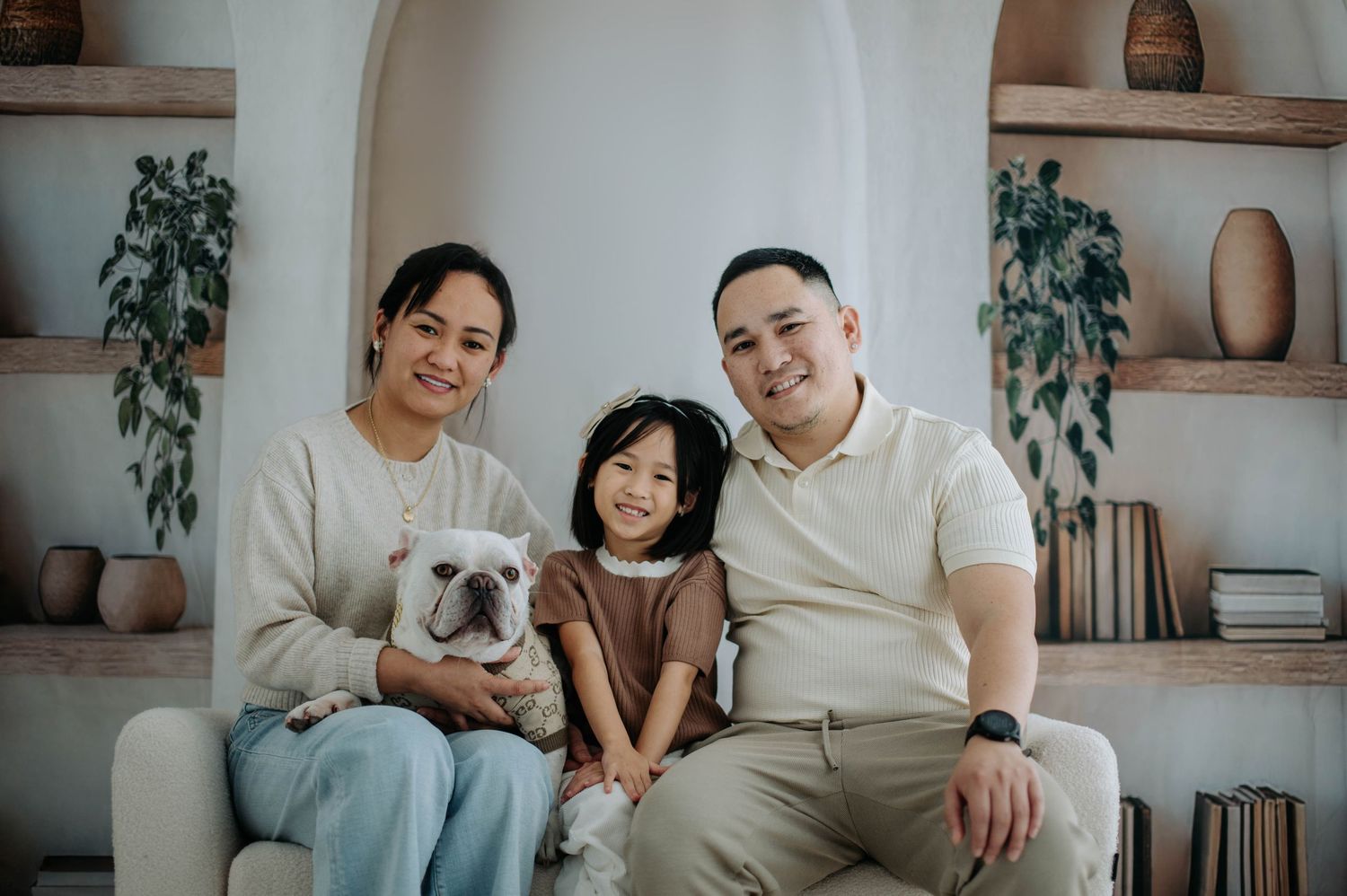 A cozy family portrait on a white sofa with their small white dog, surrounded by potted plants and decorative shelving.