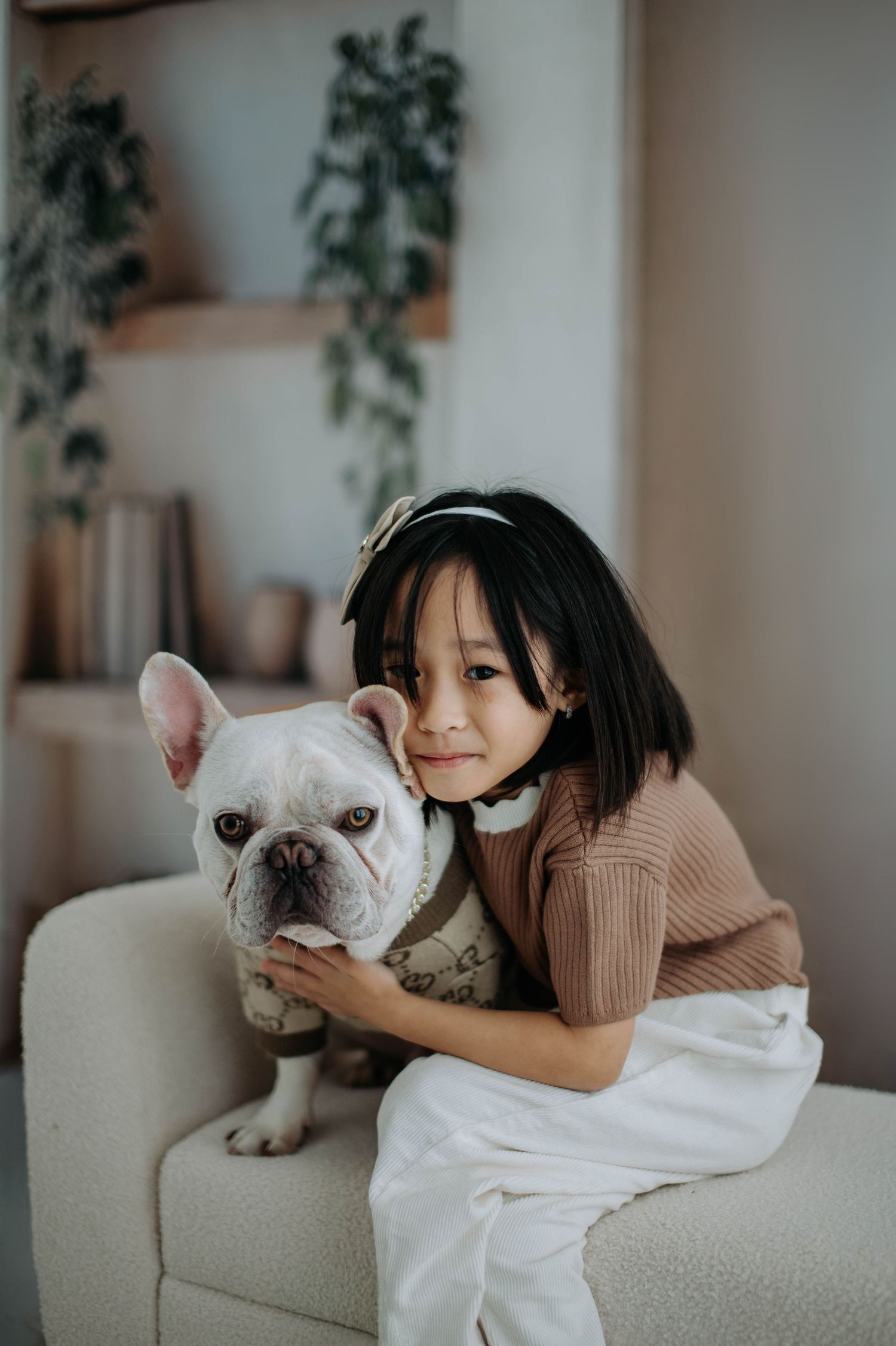 French Bulldog and companion share affectionate moment on beige couch with houseplants in background.