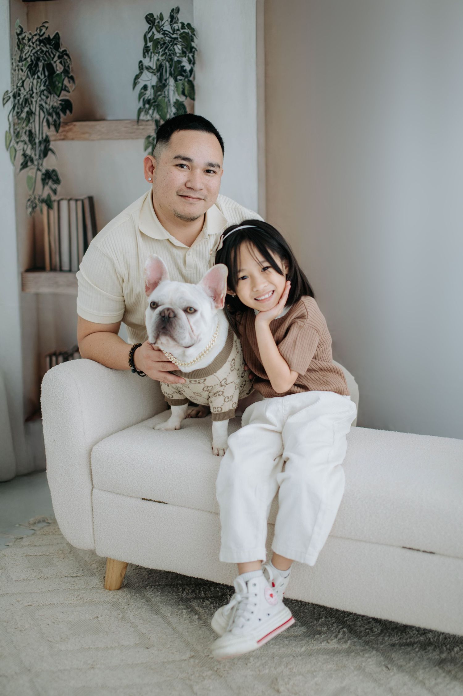A couple sits on a light gray couch with their French Bulldog in a minimalist room with potted plants.