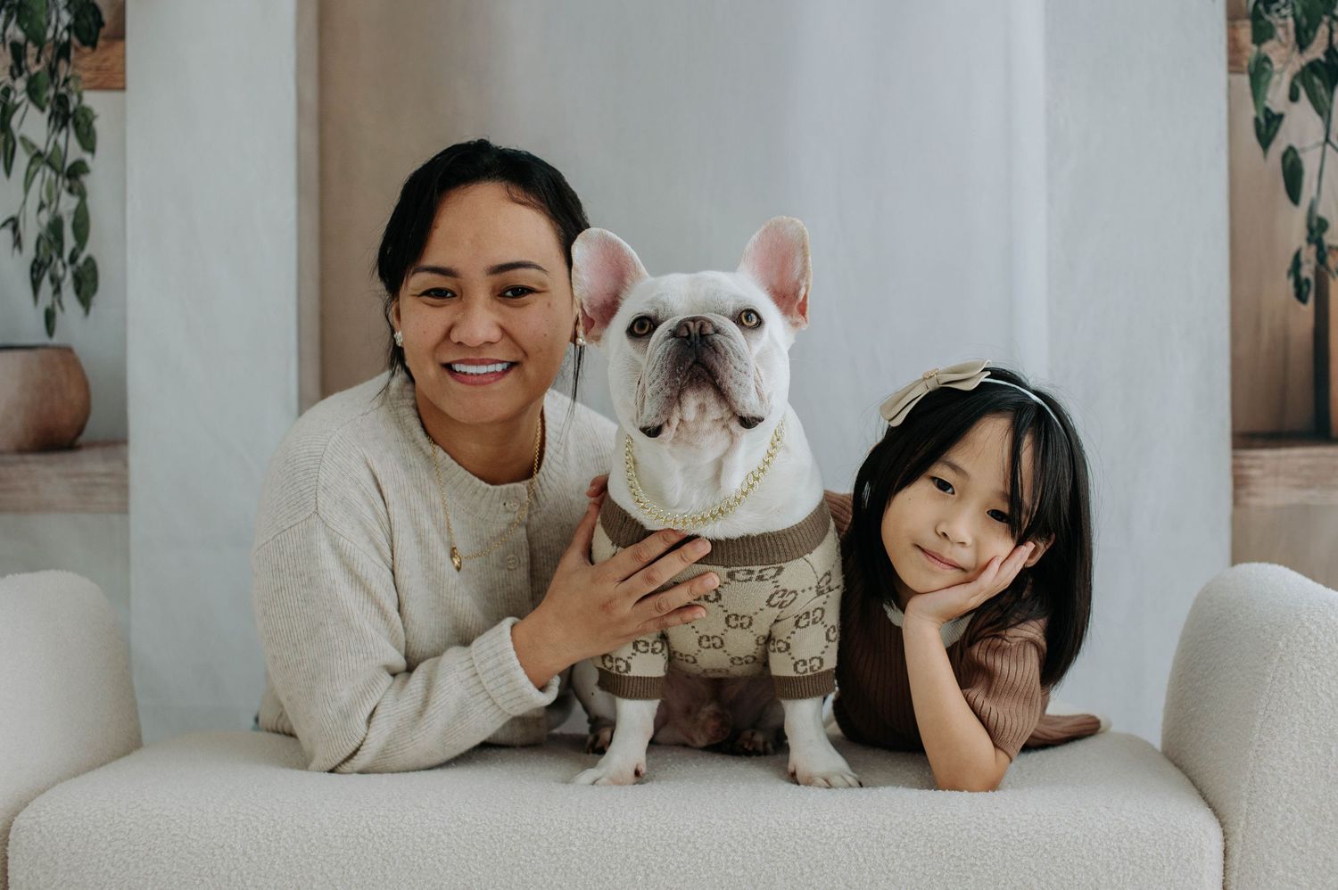 Two people sitting on a white couch with a French Bulldog wearing a patterned sweater.
