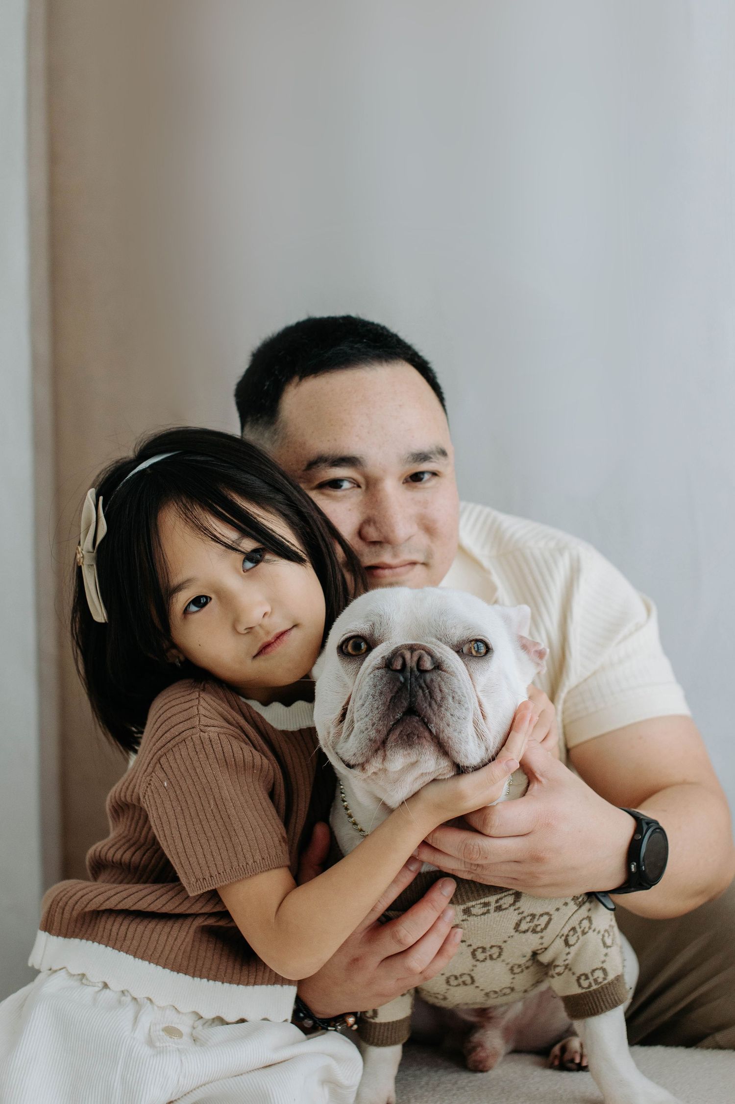 A family poses with their adorable French Bulldog wearing a designer outfit in a minimalist indoor setting.