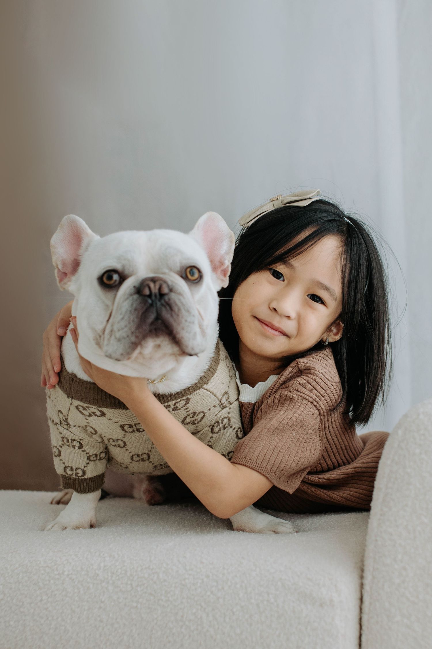 A French bulldog lounges on a light colored couch while being cuddled and hugged.