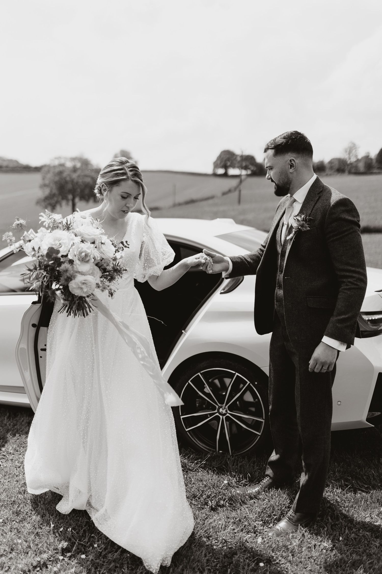 Black and white wedding photograph of bride arriving at wedding beside luxury car in countryside setting with floral bouquet.