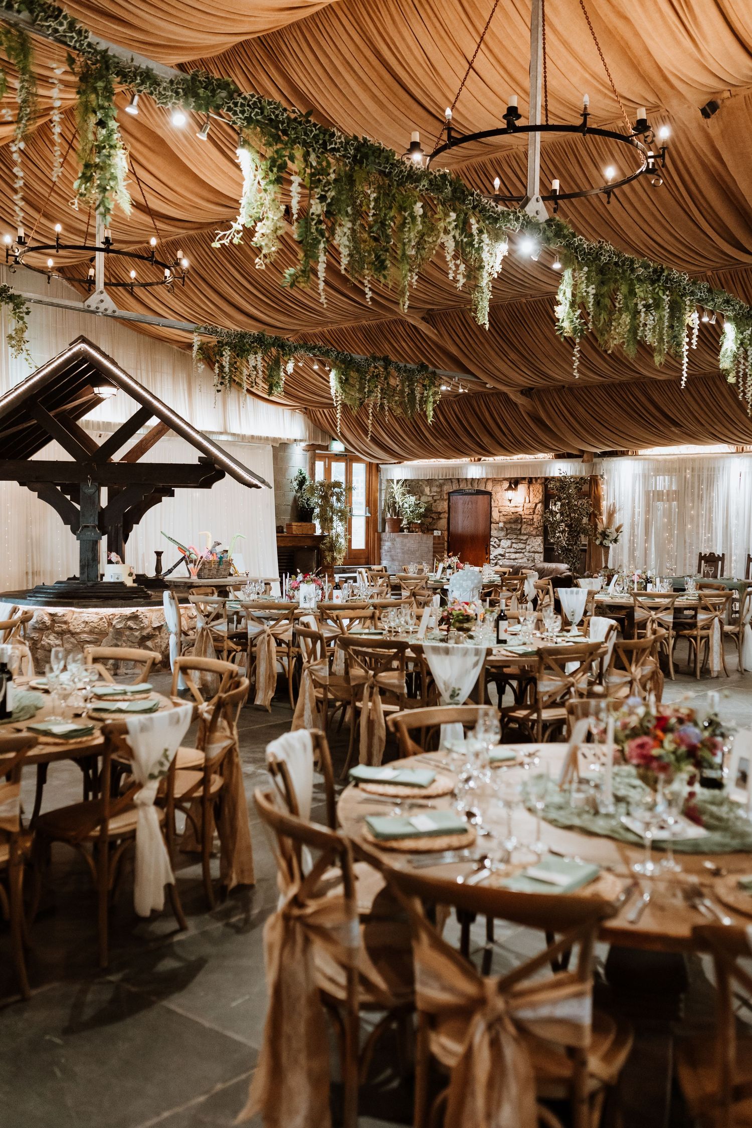 Rustic wedding reception venue with draped fabric ceiling, hanging greenery, chandeliers and wooden cross-back chairs.