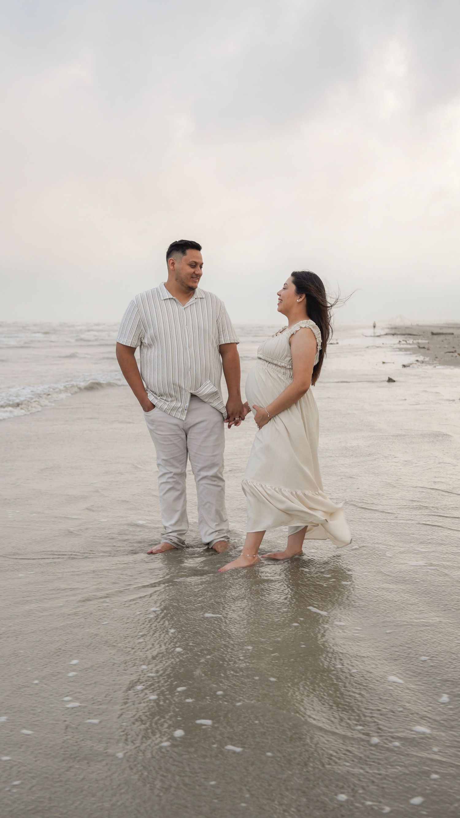 Two people holding hands and laughing while standing in the shallow ocean waves at the beach on a cloudy day.