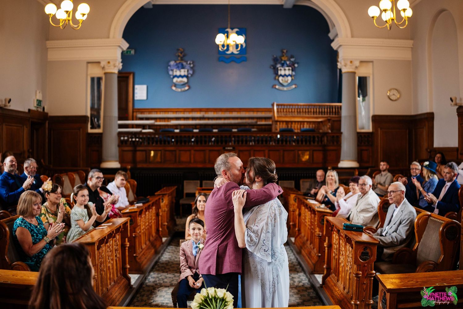 A wedding ceremony in Southport Town Hall with ornate chandeliers and pews filled with guests.