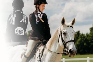 An equestrian rider in black helmet and jacket competes in a show jumping event on a white horse.