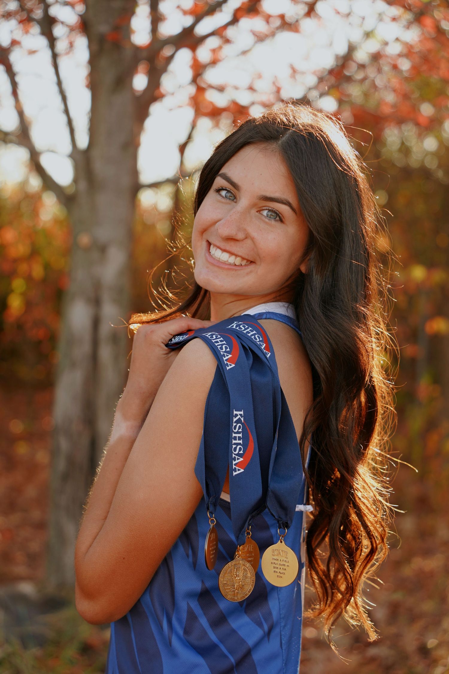Brunette teen girl with blue eyes displaying KSHSAA medals in blue athletic top among fall foliage.