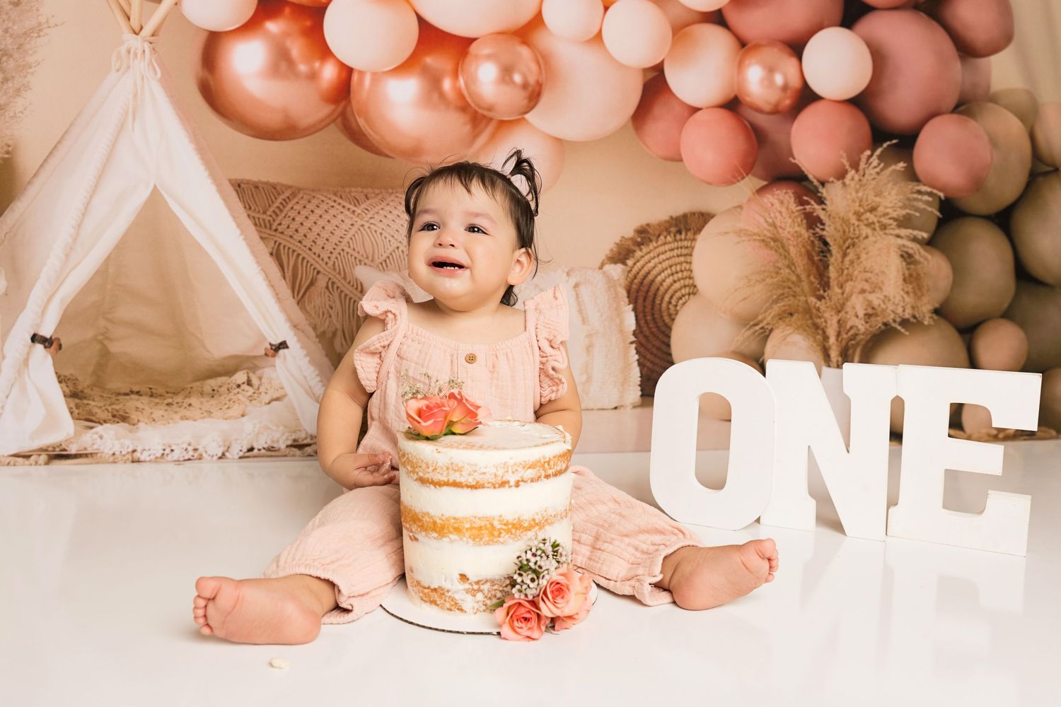 First birthday cake smash photo setup with peach balloons, teepee tent, and large white 'ONE' letters against neutral backdrop.