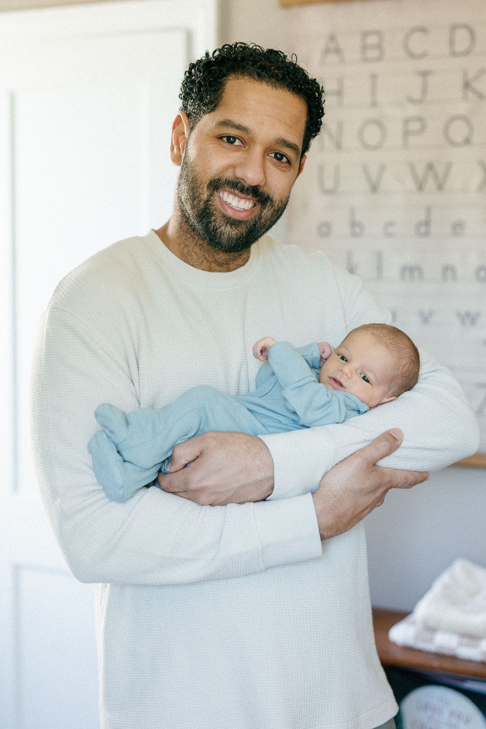A parent in a white sweater holds their newborn baby wearing light blue in front of an alphabet wall decoration.