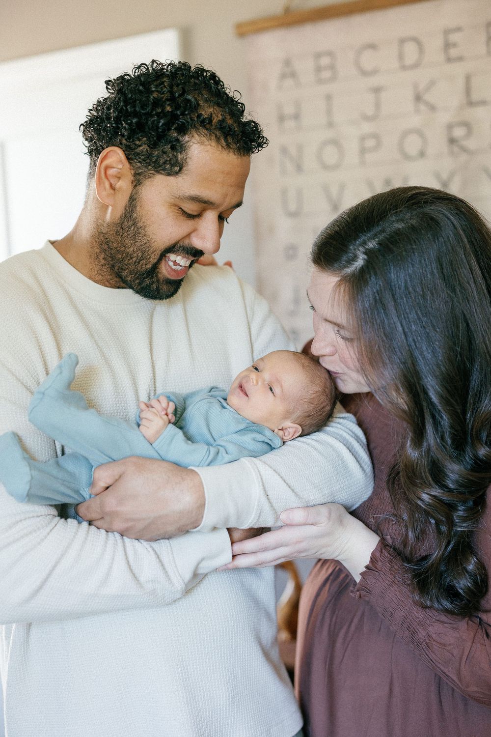 Family lovingly holds and snuggles with their newborn baby in studio setting with alphabet background on wall.