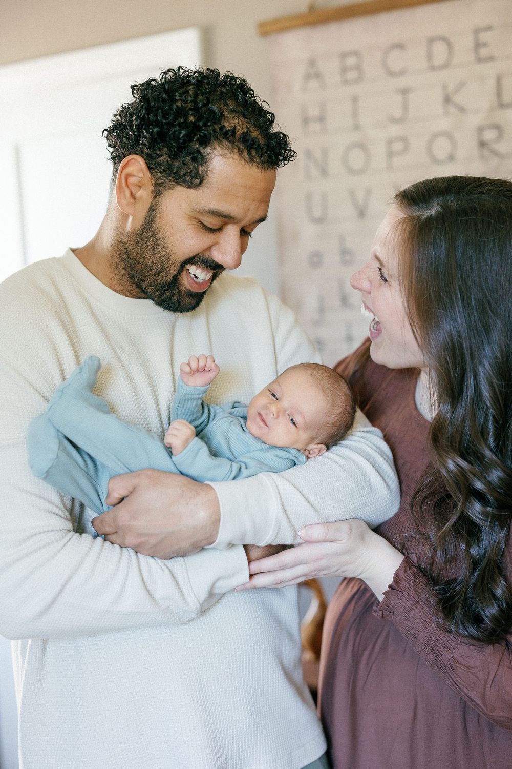 Parents sharing a joyful moment with their sleeping newborn baby in front of an alphabet wall.