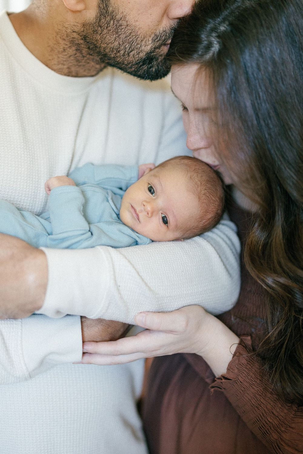 A sweet newborn baby in light blue clothing is held tenderly by parents in a close family portrait moment.