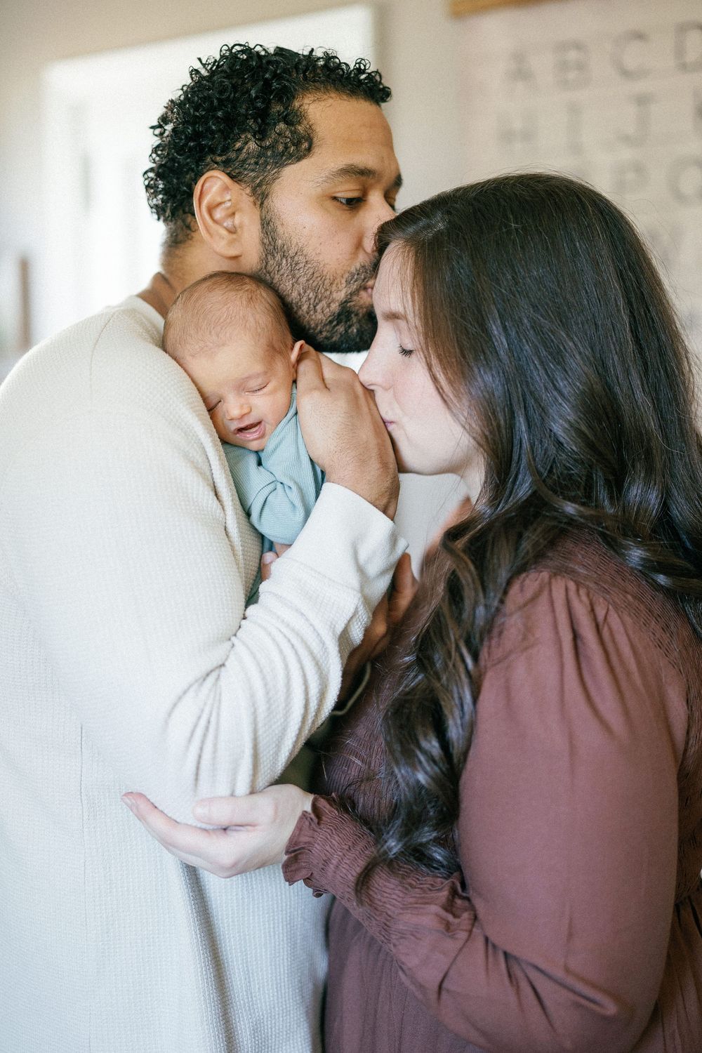 Parents tenderly embrace their sleeping newborn baby while wearing cozy sweaters at home.