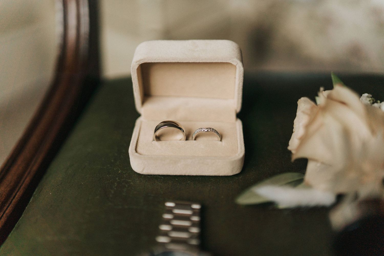 Two wedding rings nestled in a cream colored velvet box next to a white rose on a dark surface.
