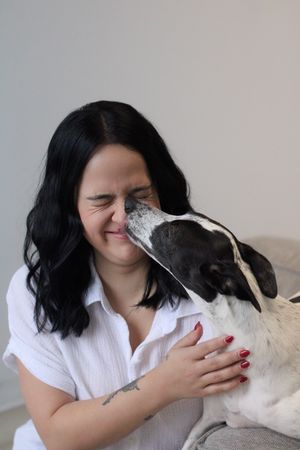 A black and white dog gives affectionate kisses to someone wearing a white shirt while sitting on a couch.