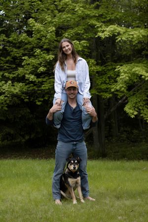 A playful outdoor portrait with two people and their black and tan dog standing in a grassy field with green trees.
