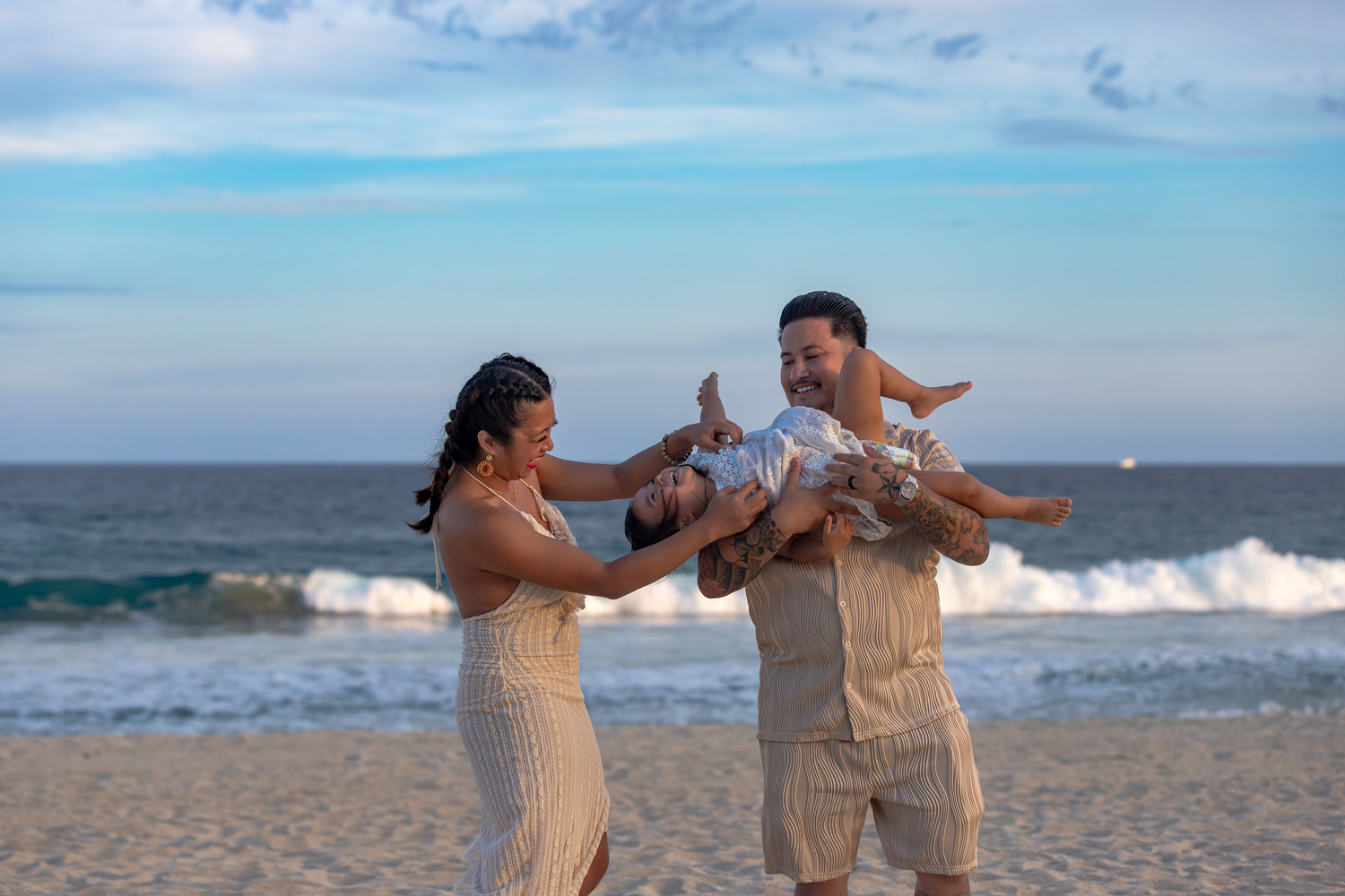 A family in white outfits poses together on a scenic beach with turquoise water and mountains in the background.