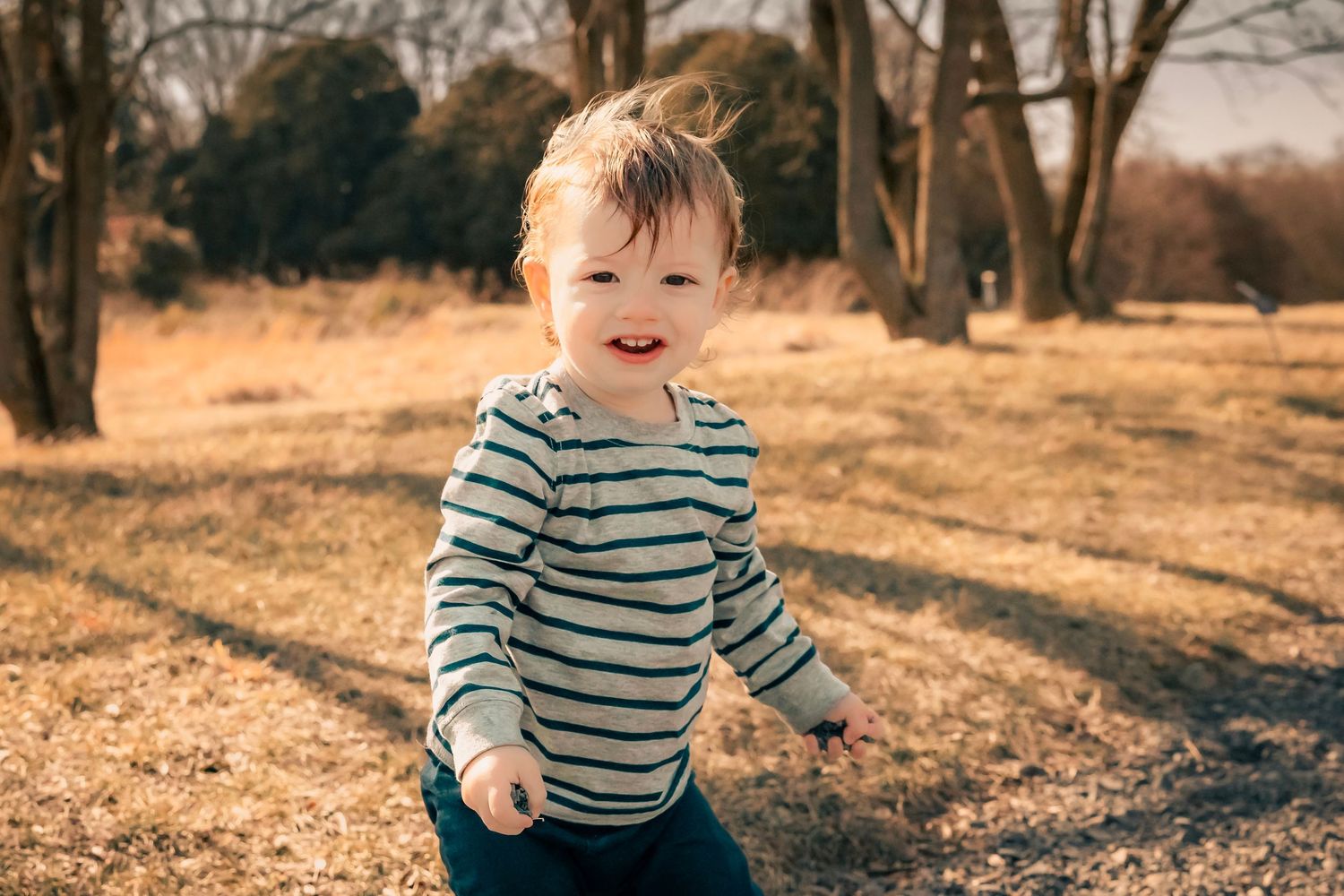 A young child wearing a striped shirt runs playfully through a golden autumn field with bare trees in the background.