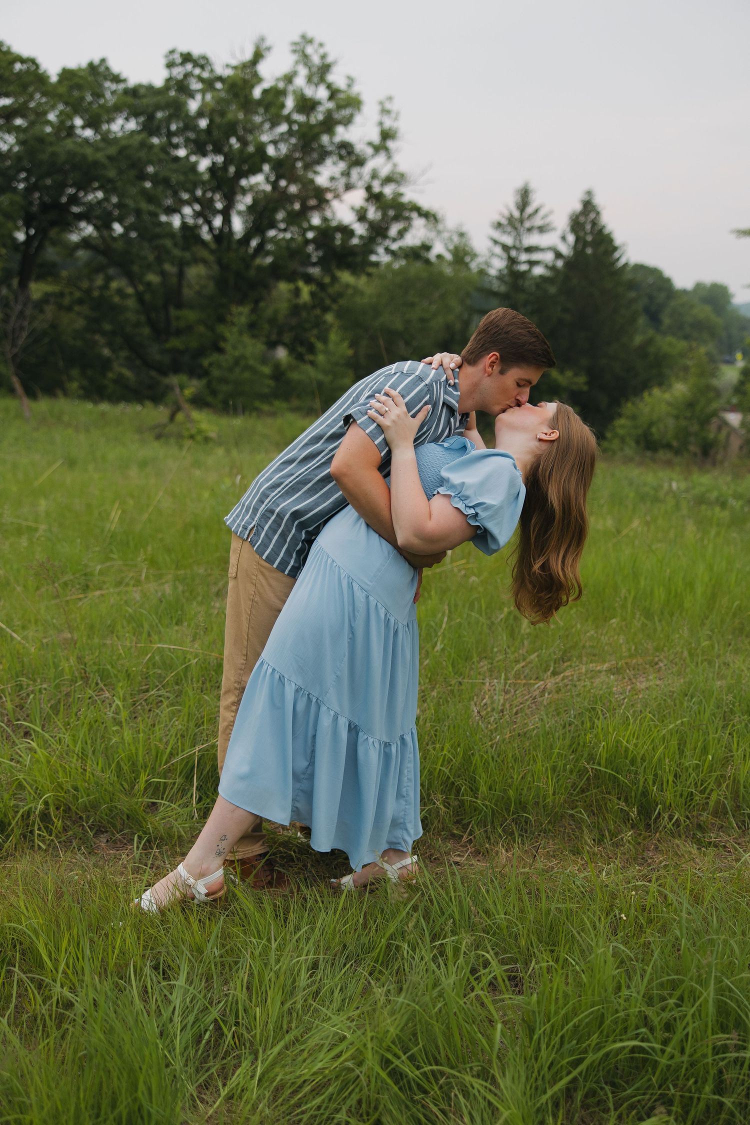 A couple in a grassy field embrace in a romantic dance-like dip pose against a natural outdoor backdrop.