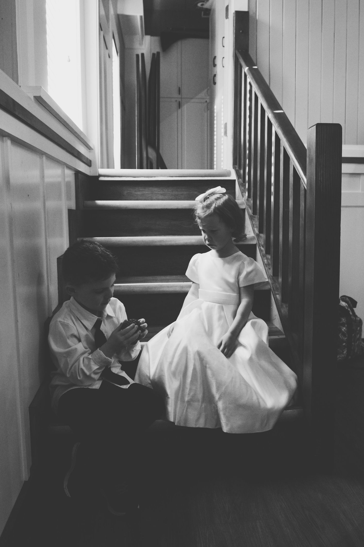 A black and white photograph of two young children in formal white attire sitting on a staircase indoors.