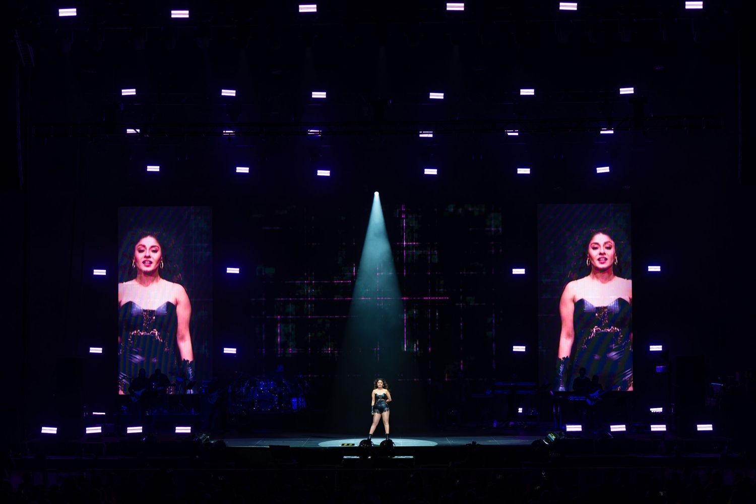 Stage performance with large video screens displaying performers and dramatic blue lighting effects in a dark theater.