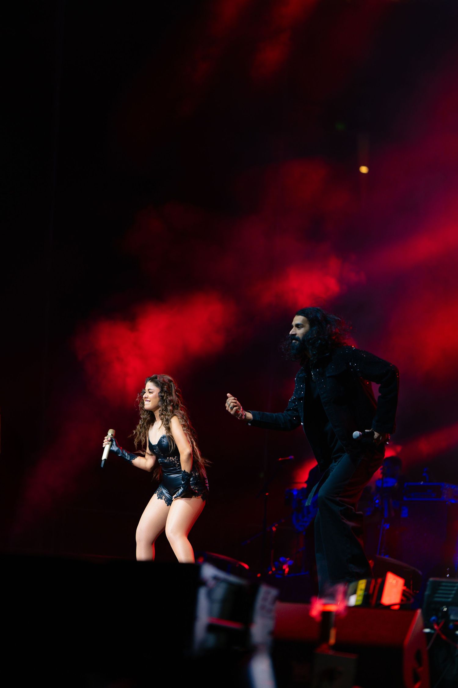 Performers dance energetically on a dark stage illuminated by dramatic red lighting and smoke effects during a concert.