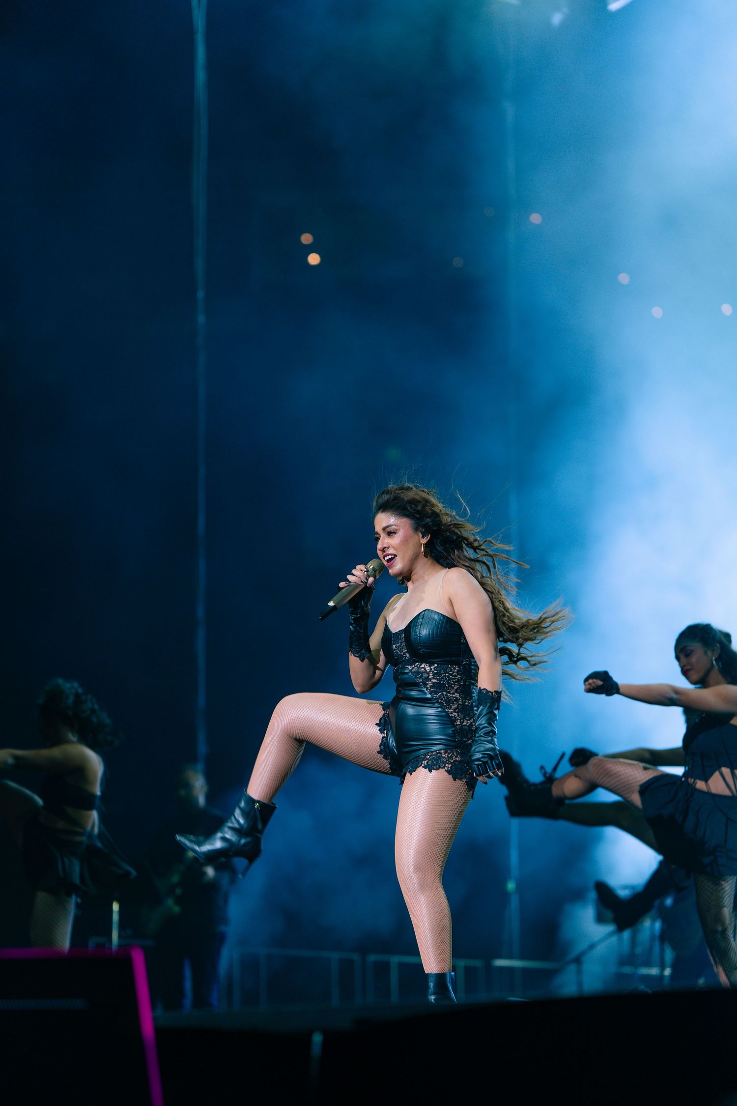 A performer in a black leotard outfit dances energetically on stage with backup dancers amid blue lighting effects.