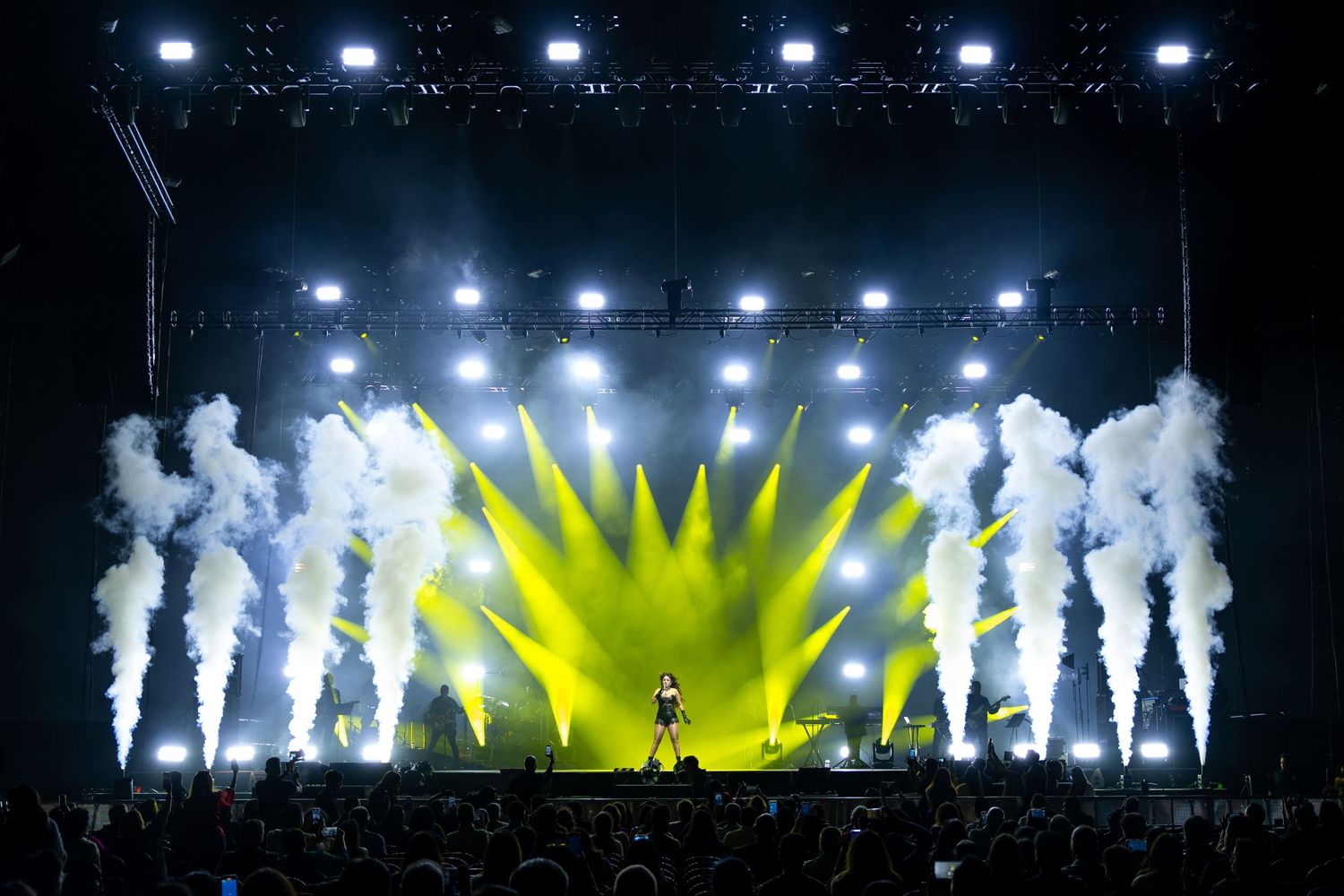 Concert stage with silhouetted performers against bright yellow and white spotlights creating a dramatic backlit effect.