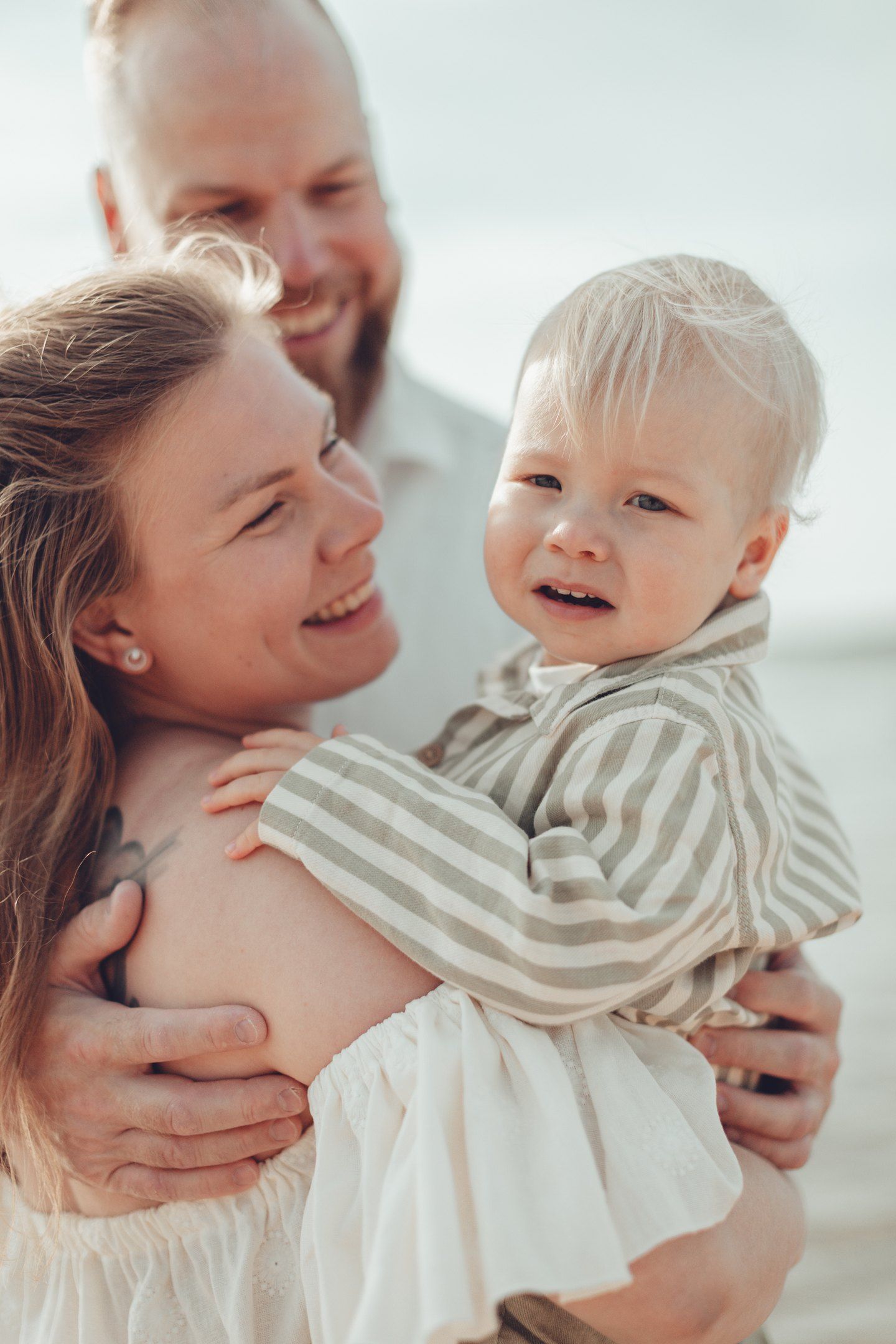 Happy family with smiling parents holding their blonde toddler outdoors near water on a sunny day.