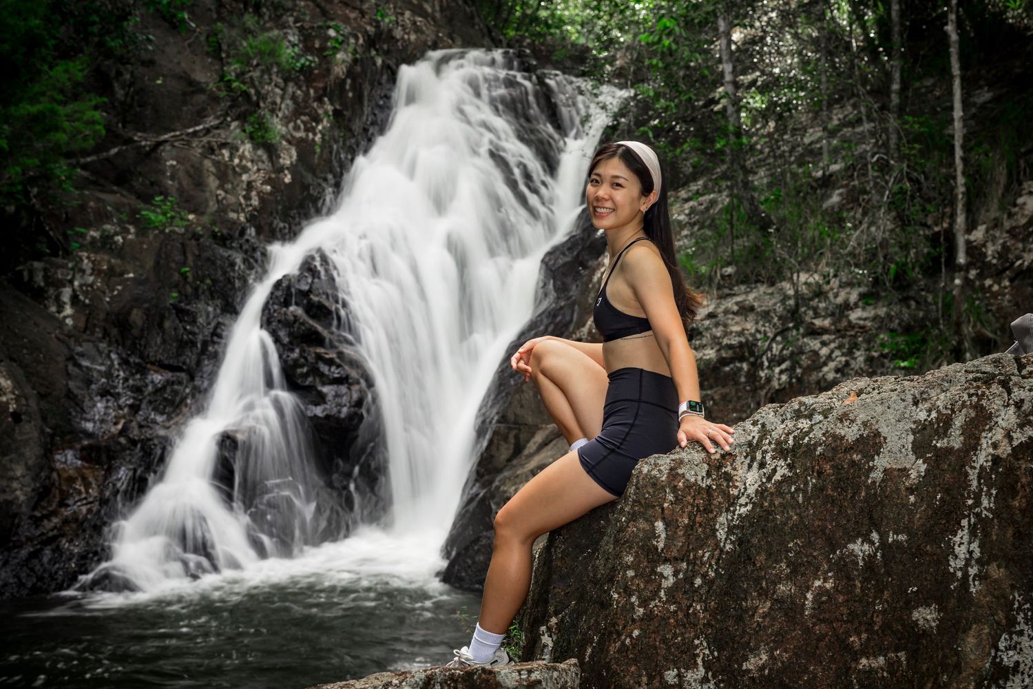 A hiker in athletic wear poses on rock formations beside a cascading waterfall in a lush forest setting.