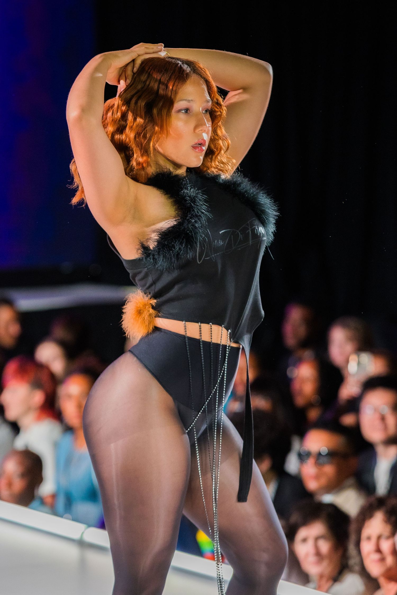 Performer in black leotard and tights strikes dramatic pose on fashion runway with audience in background.