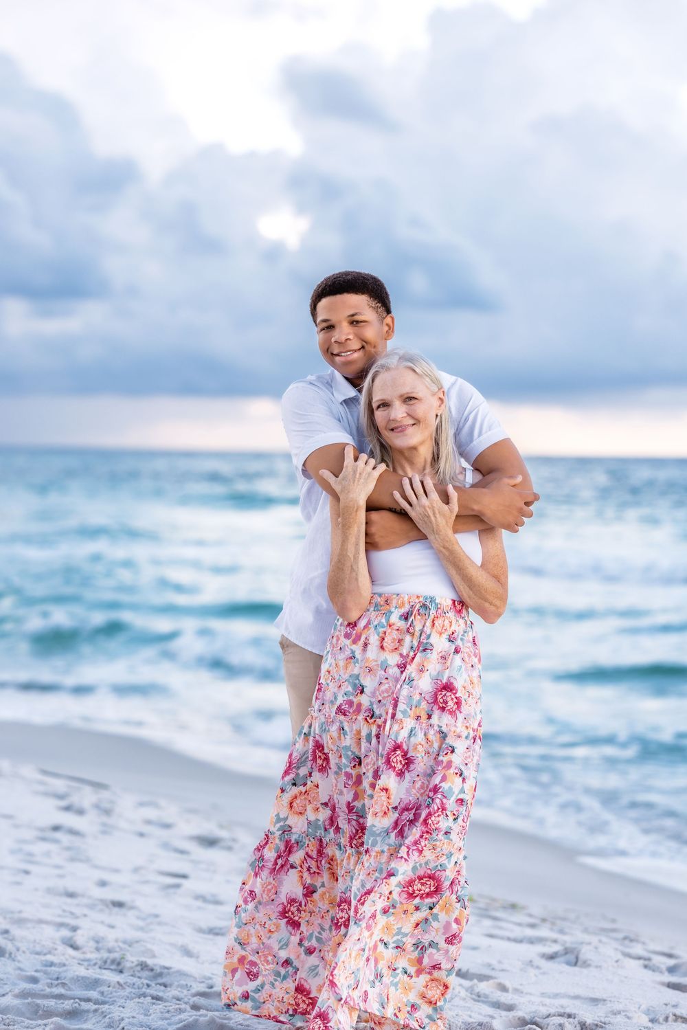 Romantic couple embraces on beach at sunset with waves and cloudy sky in background.