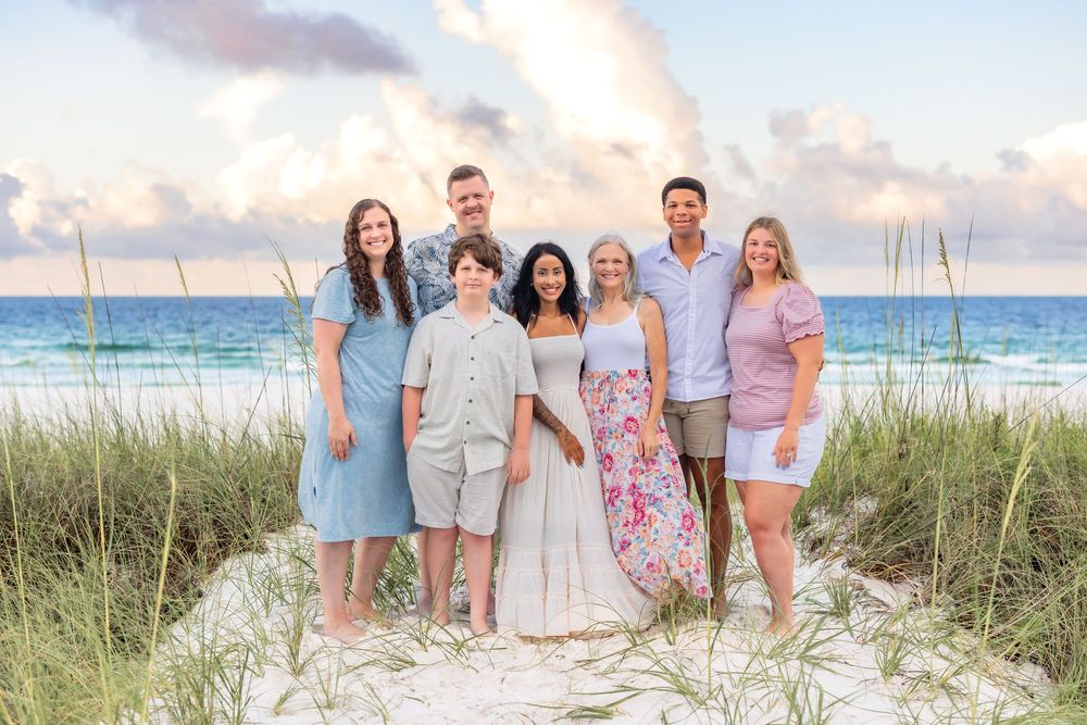 A group dressed in soft pastel colors poses together on a sandy dune with ocean and sunset sky in background.