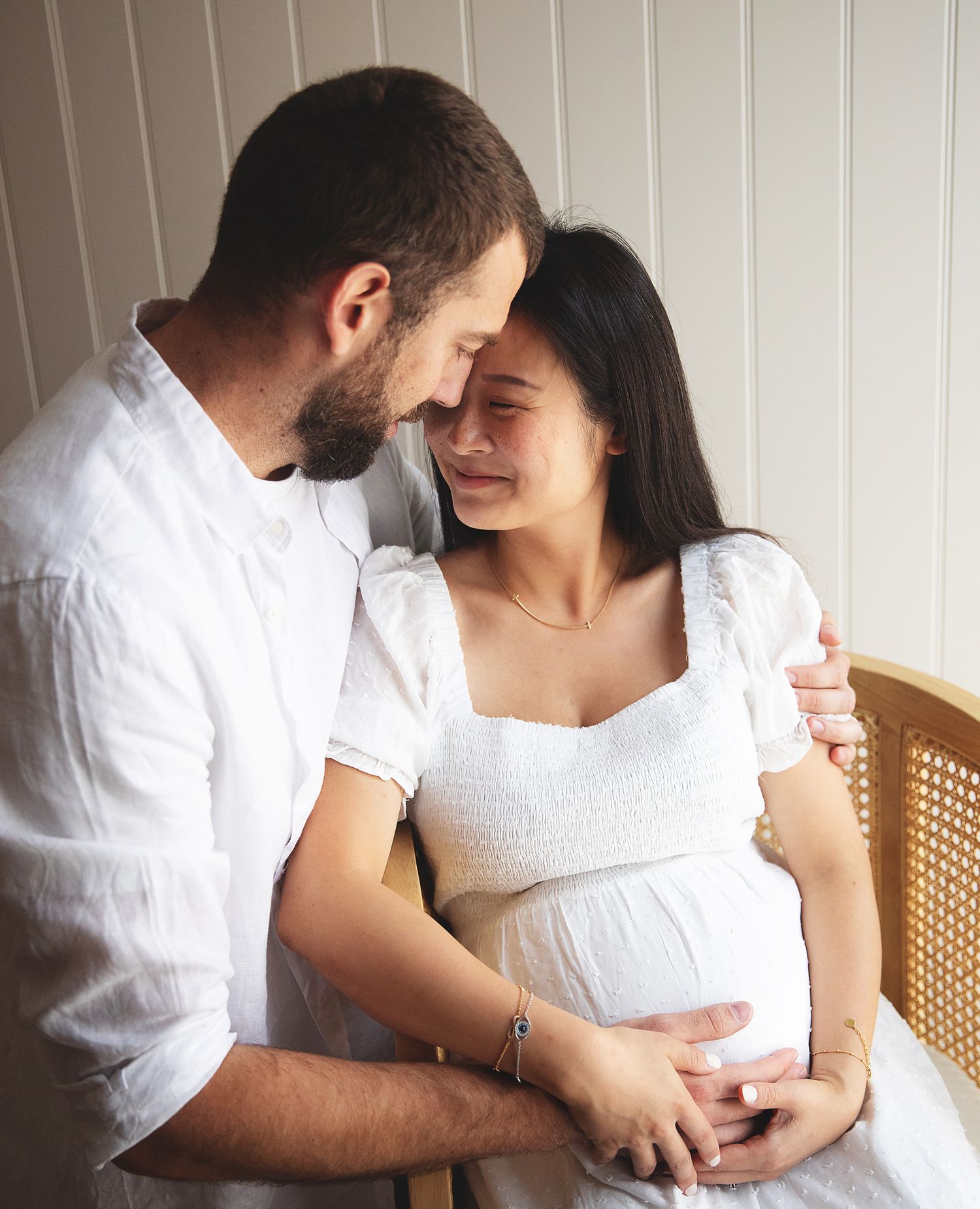 Expecting couple in white clothing share an intimate moment as they caress her pregnant belly by a white wall. During a Bexley photoshoot