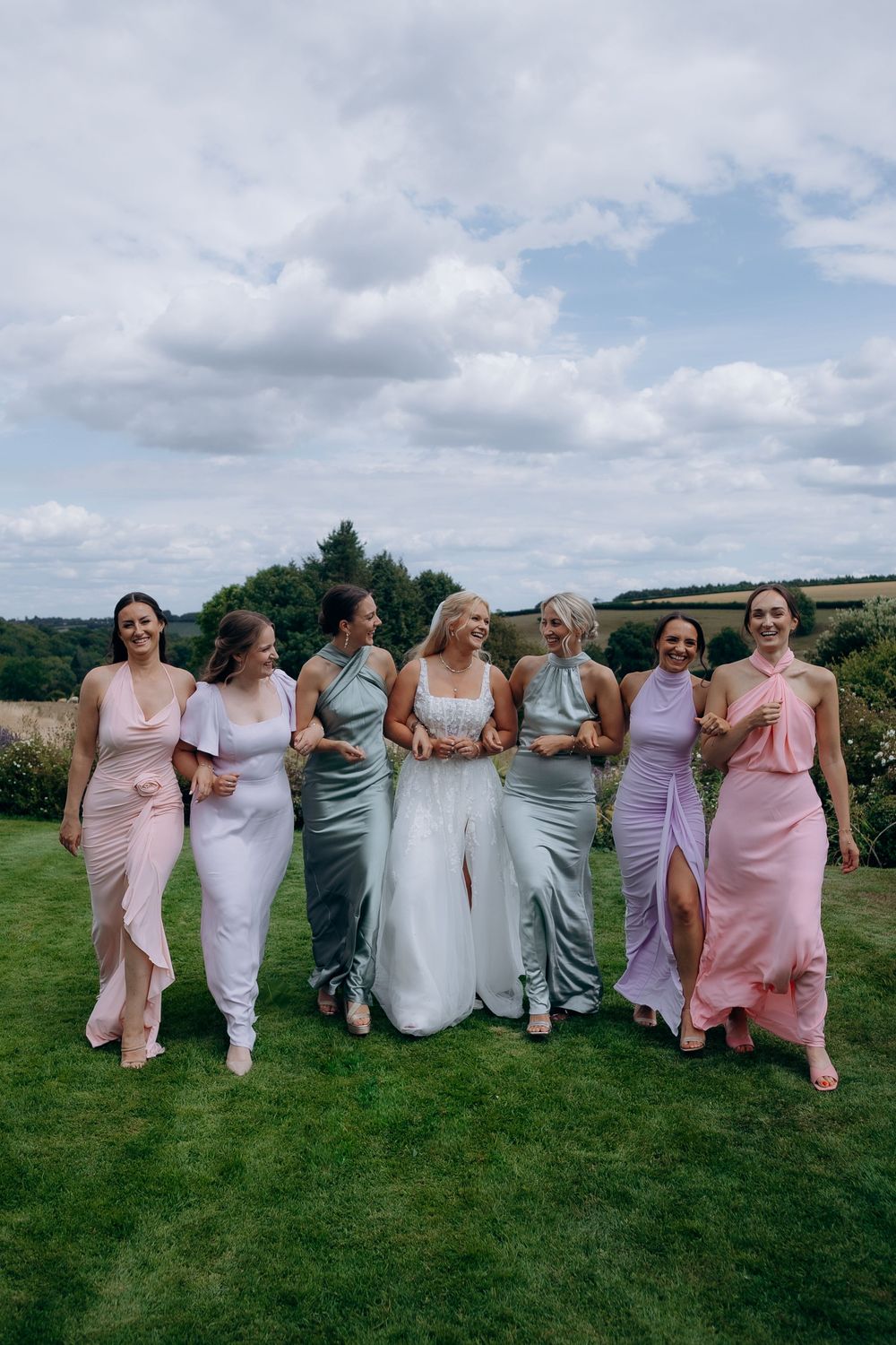 Wedding party in pink, purple and sage dresses posing in a line outdoors on green grass.