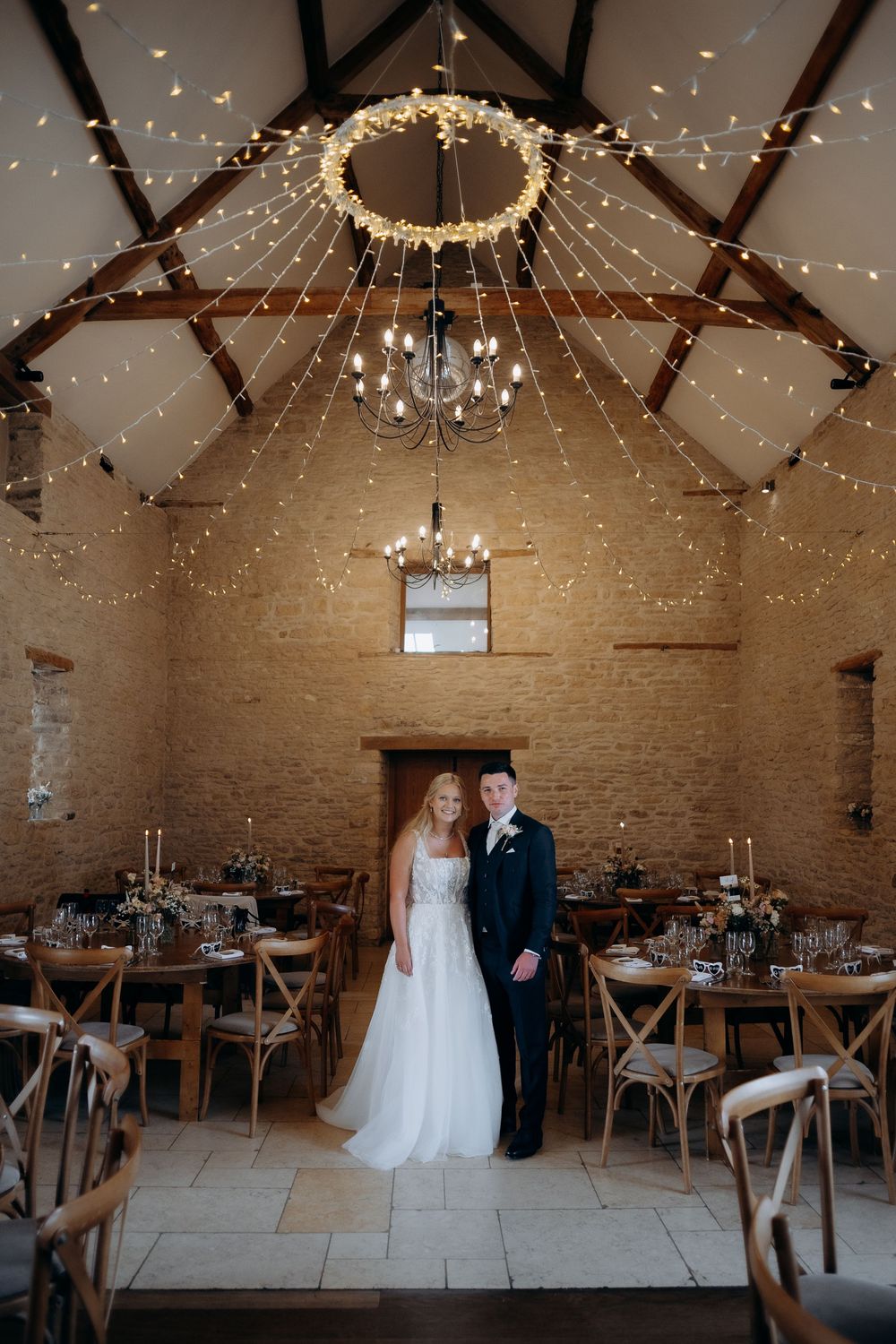 Rustic barn wedding venue interior with exposed beams, string lights and chandeliers creating romantic ambiance for reception.