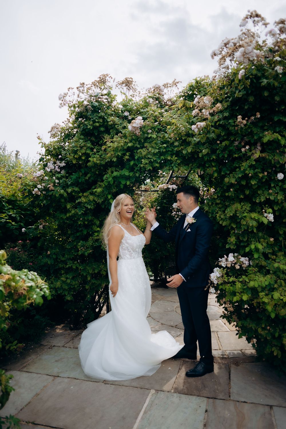 A newlywed couple shares a romantic moment under flowering rose bushes in an outdoor garden setting.