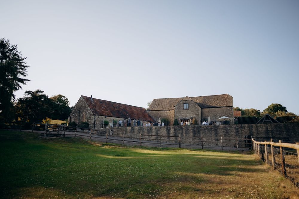 Stone farmhouse wedding venue with manicured lawn and wooden fence under blue summer sky.
