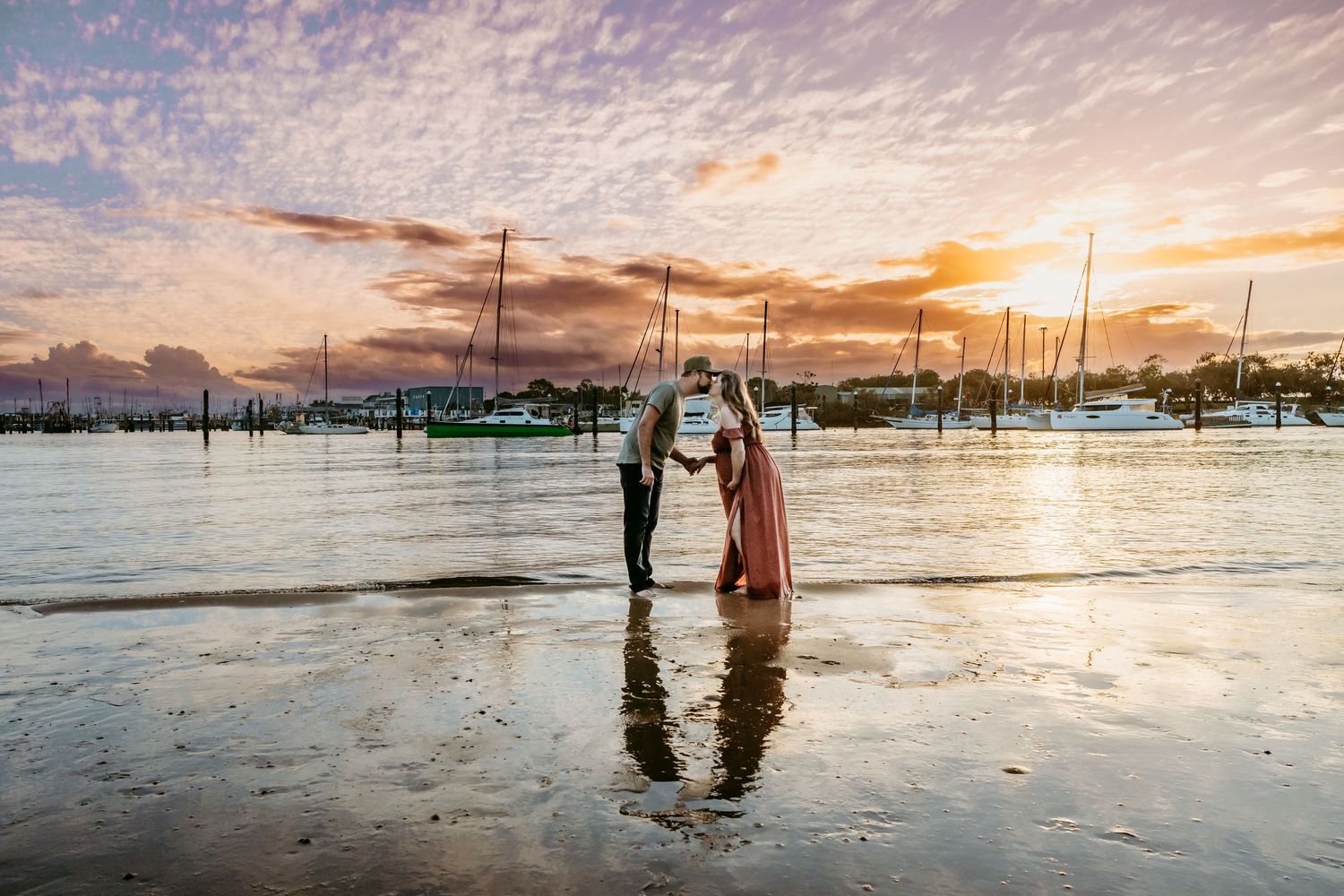 Two people stand together at sunset on a tidal beach with boats and docks in the background.