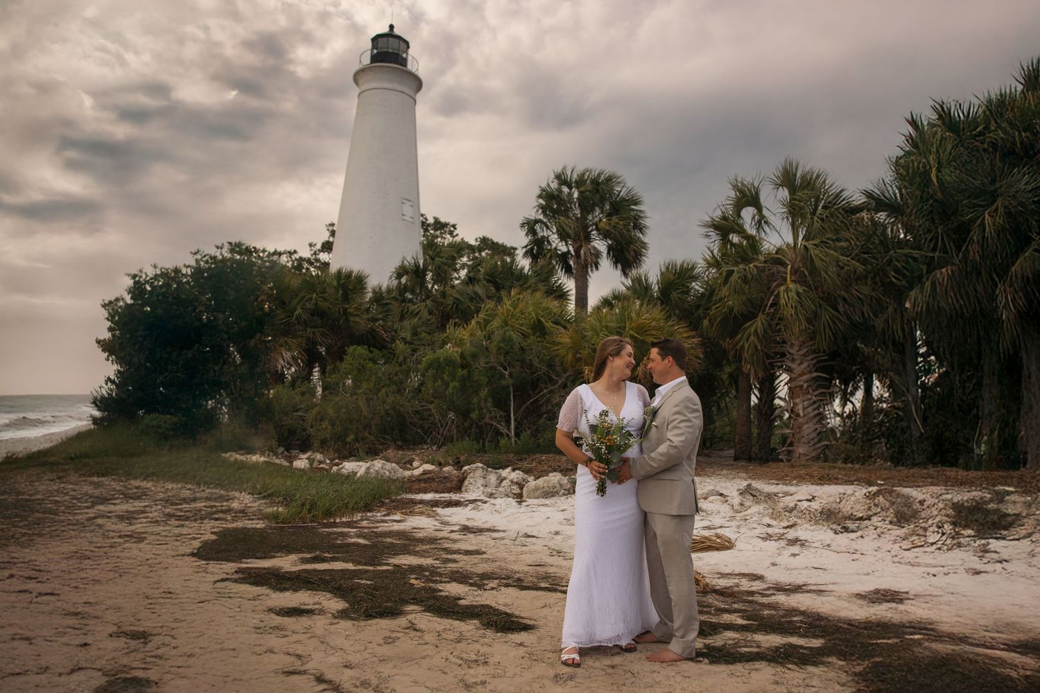 A romantic wedding couple embraces on a sandy beach with the historical St. Marks lighthouse and palm trees in the background.