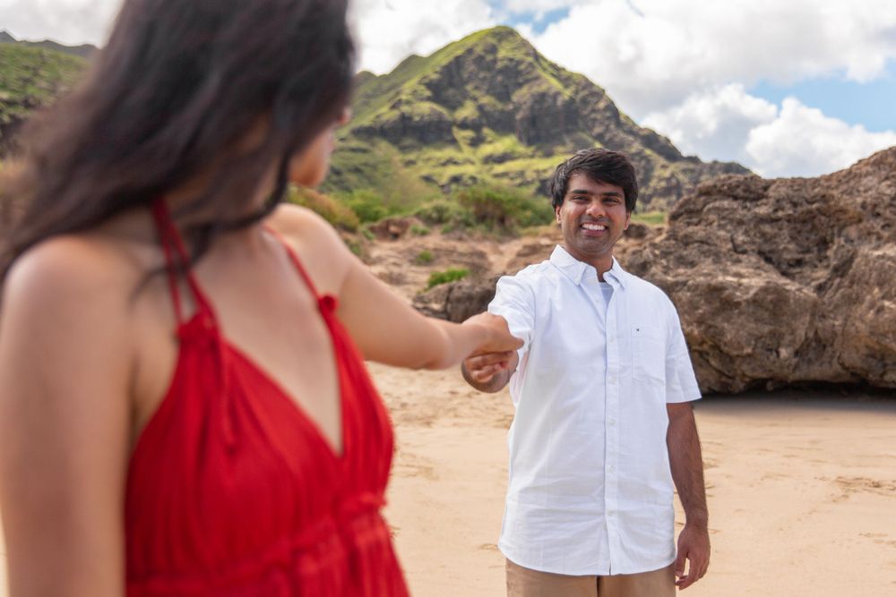 A couple shares a romantic moment on a sandy beach with dramatic mountains and rock formations in the background of Makua Beach.
