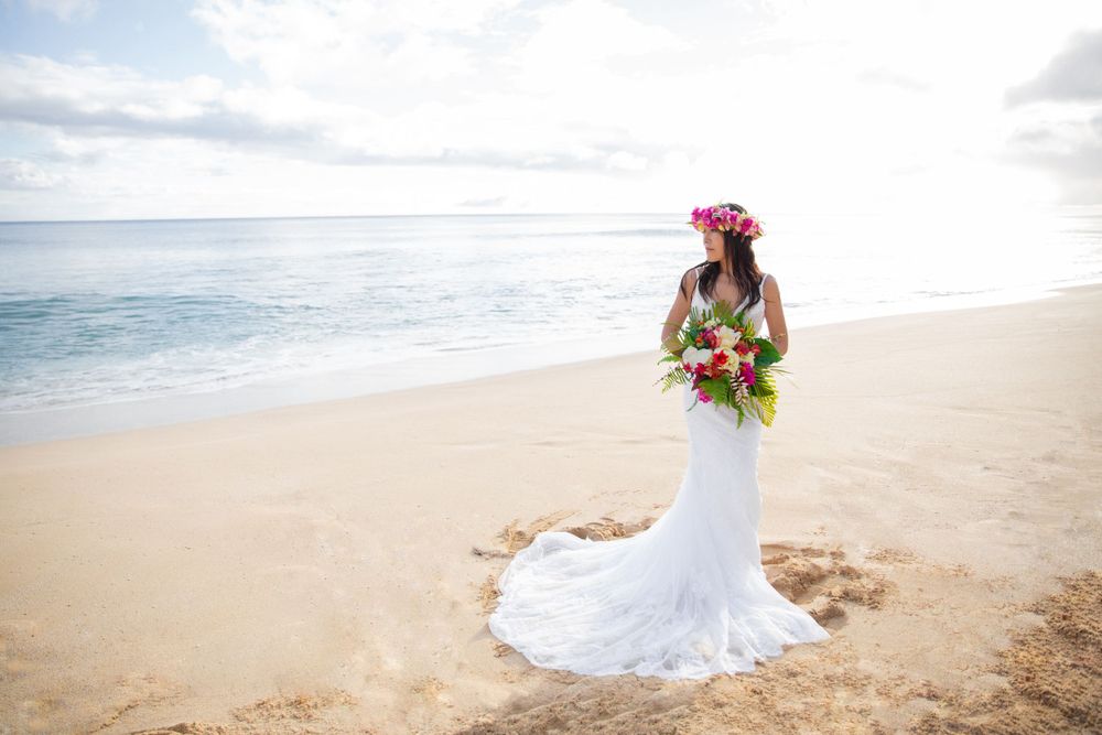 A bride in a white lace gown and flower crown holds a tropical bouquet while standing on Yokohama Beach at sunset.