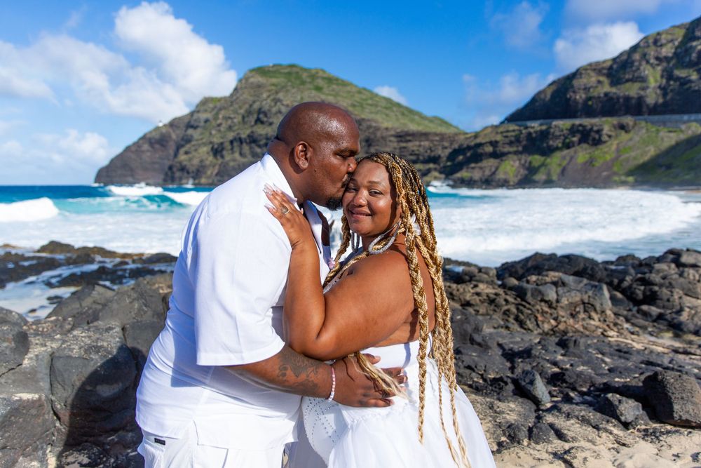 A couple in white attire embrace on a rocky beach with dramatic cliffs and crashing waves at Makapuu Beach Park.