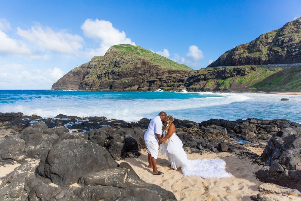 A couple in wedding attire embraces on volcanic rocks along scenic Makapuu Beach Park with turquoise waters and mountains.